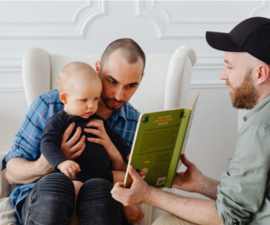 Two men sitting and reading a book to a baby who is held by one of the men.