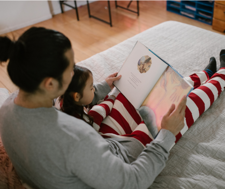 A person reading a book to a child while lying on a bed. Both are wearing pajamas.